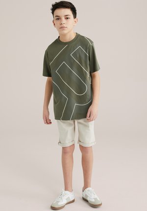Grünes T-Shirt mit kurzen Ärmeln, das ein großes weißes Logo-Design zeigt. Kombiniert mit hochgekrempelten beigen Shorts und weißen Sneakers mit grünen Akzenten.