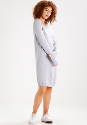 Robe pull grise en tricot, à manches longues avec des poignets côtelés, encolure dégagée, longueur mi-cuisse, portée avec des baskets blanches. Texture lisse.