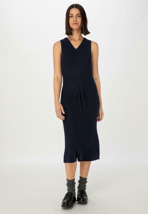 Ärmelloses, knielanges Kleid aus navy-blauem Rippstrickstoff mit V-Ausschnitt. Besitzt eine Kordelzugtaille und einen Schlitzsaum, kombiniert mit grauen Socken und schwarzen Schuhen.