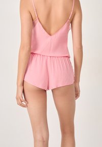 Modelka ubrana w różowy, bezrękawnikowy kombinezon z niskim tyłem i koronkowymi detalami, zwrócona tyłem do kamery. Delikatne, neutralne tło.