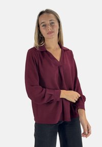 Bordeaux blouse met een kraag, lange mouwen, een gladde textuur en een losse pasvorm, gecombineerd met zwarte jeans.