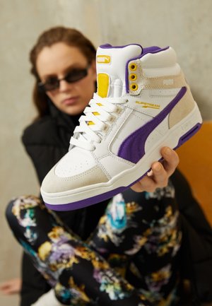 Osoba trzyma białego, wysokiego sneakersa Puma z fioletowymi i żółtymi detalami, skupiając się na bucie w swobodnym, wewnętrznym otoczeniu.