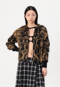 Chaqueta bomber de punto con estampado de leopardo en marrón y negro, con capucha y puños acanalados. Llevada sobre un top corto negro y una falda de cuadros.
