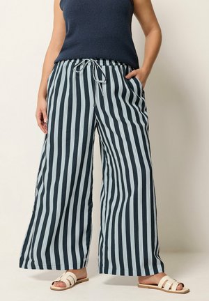 Kobieta nosząca szerokie spodnie w pionowe, czarno-białe paski z wiązaniem w talii oraz białe, płaskie sandały z odkrytymi palcami.