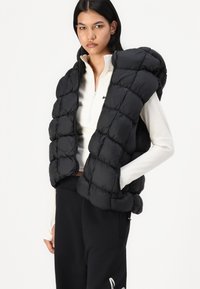 Melnā pūkainā veste ar quiltotu dizainu, ar augstu apkakli un atvērtu priekšpusi. Valkāta virs baltas ar rāvējslēdzēju aizvērtas augšdaļas ar garām piedurknēm.