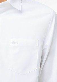 Witte langlopende shirt met geknopte kraag, voorzien van een borstzak en een subtiele geborduurde logo. Vloerzijde stof met een strak ontwerp.