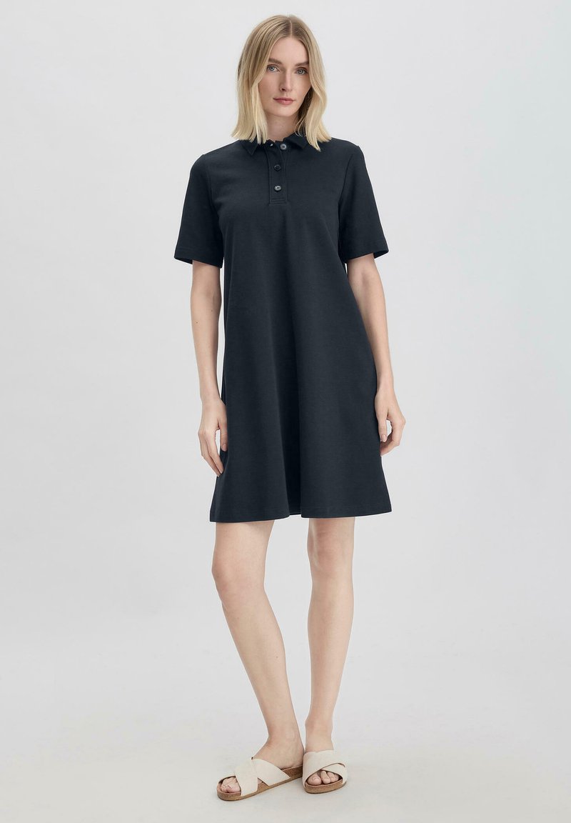 Schwarzes Polokleid mit kurzen Ärmeln, geradem Schnitt und dreiknöpfiger Knopfleiste. Glattes Baumwollgewebe, knielang, kombiniert mit hellen Sandalen.