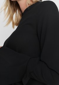 Sort bluse med lange, udflydende ærmer og høj krave. Glat stof med en subtil glans og bløde flæser ved ærmekanterne.