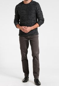 Pull en maille texturée noire à manches longues, associé à un pantalon chino gris foncé près du corps et des bottines noires, présenté sur un fond blanc.