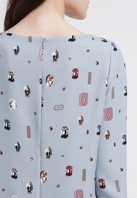 Jasnoniebieska tkanina z rozrzuconymi kolorowymi wzorami kamieni szlachetnych i teksturowymi akcentami, z prostym okrągłym dekoltem i dyskretnym zapięciem na plecach.