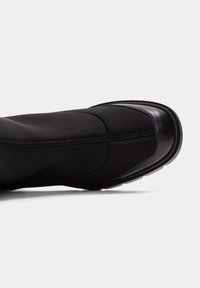 Bota negra que presenta una combinación de tela suave y cuero liso, con puntera redondeada, suela de goma gruesa y costuras detalladas.