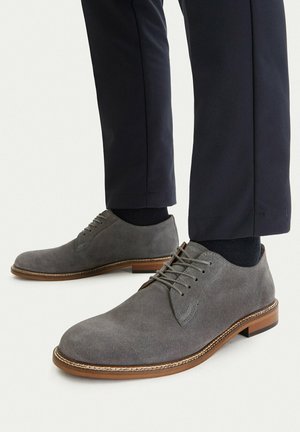 Man wearing black pants and gray suede lace-up dress shoes with brown soles, standing on a white background.