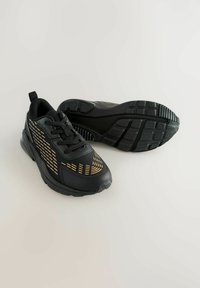 Schwarze Sportschuhe mit einer glatten Textur, die goldene Akzente in einem Zickzack-Muster aufweisen und eine gepolsterte Sohle für Komfort haben.