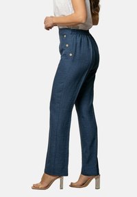 Pantalones azules de cintura alta hechos de un tejido texturizado, con un corte relajado, bolsillos laterales y detalles de botones a lo largo de la cintura. Tacones transparentes de bloque.