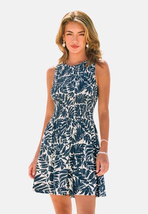 Frau mit hellbraunen Haaren, die ein ärmelloses weißes Kleid mit dunkelblauen tropischen Blattmustern trägt, dazu Ohrringe und Armbänder.