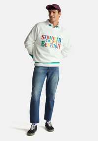 Felpa bianca con scritta multicolore "STRANGER COLORS OF BENETTON", abbinata a jeans denim blu e scarpe da ginnastica nere con lacci bianchi.