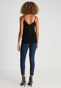 Kvinna med lockigt hår som bär en svart ärmlös topp, blå tajta jeans och metalliska ankelboots, stående vänd bort från kameran mot en enkel bakgrund.