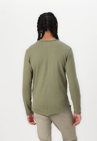 Langärmliges Hemd in olivgrün, aus weichem Stoff, mit abgerundetem Saum, sichtbaren Nähdetails und minimalistischer Gestaltung.