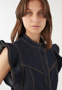 Dunkelblaue Jeansweste mit gepufften Ärmeln, die gelbe Nähte, einen hohen Kragen und ausgeprägte Paneeldetails aufweist und ein strukturiertes Design präsentiert.