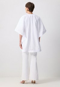 Bílá oversize tunika s krátkými rukávy, texturovaná tkanina, ve spojení s bílými rozšířenými kalhotami a sandály na vysokém podpatku, z pohledu zezadu.