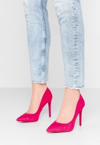 Escarpins en suède rose avec un bout pointu et un talon aiguille. Ce modèle présente un décolleté bas et une texture lisse. Portés avec un jean délavé.