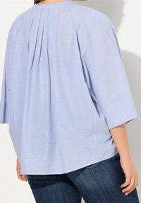 Lichtblauwe blouse met een losse pasvorm, driekwart mouwen, plooidetails aan de achterkant en een gladde stoftextuur.