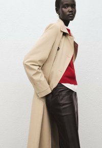 Beżowy płaszcz trencz z dwurzędowym zapięciem, noszony na czerwonym swetrze i białej bluzce, w połączeniu z ciemnobrązowymi skórzanymi spodniami.