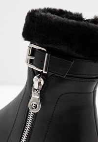 Bottine en cuir noir avec un revers doux en fourrure noire, une boucle argentée et un détail de fermeture éclair latérale, présentant une texture lisse et un design élégant.