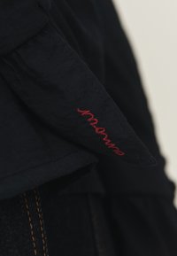 Gros plan sur une manche en tissu noir avec du texte en cursive rouge brodé et des détails de couture visibles.