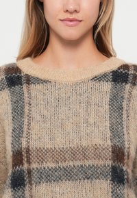 Beige gebreide trui met een geruit patroon met donkerblauwe, bruine en grijze accenten. Zachte textuur en brede halslijn.