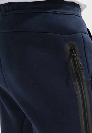 Marineblå bomuldsjoggers har en glat tekstur, elastisk taljebånd og lynlåslommer i siden med sorte detaljer.