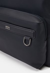 Mochila azul marino con tejido texturizado, equipada con un bolsillo con cremallera y un lazo de hardware plateado. Logo embossado en el panel frontal.