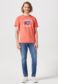 Raudona medvilninė marškinė su "WRANGLER 47 COWBOYS" grafika, apvaliu apykaklės iškirpimu, trumpomis rankovėmis, derinama su mėlynais džinsais ir baltais sportiniais bateliais.