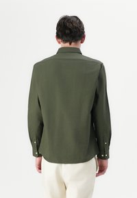 Camicia verde oliva a maniche lunghe con colletto e polsini abbottonati, caratterizzata da una texture liscia e un orlo dritto sul retro.