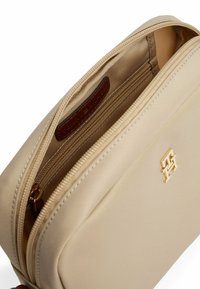 Borsa in nylon beige con una texture liscia, dotata di una cerniera dorata e di un accento con logo dorato. L'interno presenta un compartimento foderato e un'etichetta marrone.
