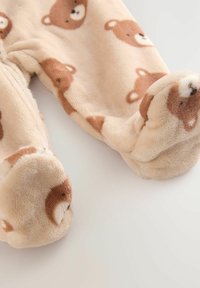 Mjuk, beige tyg med ansikten av bruna björnar pryder fötterna på en mysig spädbarnsoverall, vilket betonar värme och komfort.