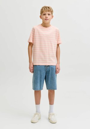 Svetlolasi fant stoji, oblečen v roza črtasto majico, modre jeans kratke hlače, bele nogavice in bež superge na enobarvnem svetlem ozadju.