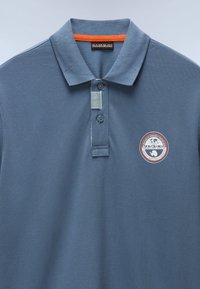 Modrá polo košile vyrobená z texturované tkaniny s tříknoflíkovou légou, oranžovým akcentem na límci a kulatou nášivkou loga na hrudi.