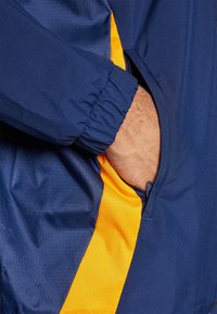 Navyblauwe jas met gele accenten. Bevat een ritszak en ribgebreide manchetten. Het materiaal lijkt glad met een subtiel textuurpatroon.
