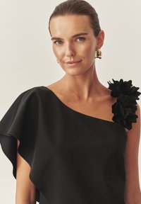 Fekete ruha egy vállra nyújtott dizájnnal, fodros ujjal és virágos díszítéssel. Simább anyag, matt felülettel.