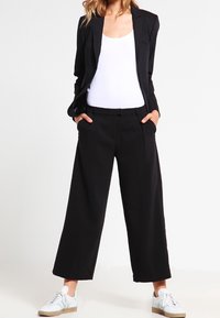 Giacca nera su misura e pantaloni a gamba larga, abbinati a una canotta bianca e sneakers. Tessuto liscio, design strutturato, linee pulite.