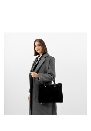 Vrouw draagt een lange grijze jas en houdt een zwarte handtas met gouden accenten vast tegen een effen witte achtergrond.