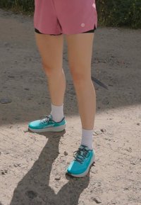 Turkise joggesko med svarte detaljer, hvite sokker, brukt med rosa treningsshorts. Fremtredende snøring og teksturert såle på grusoverflate.