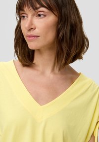 Gelbes V-Ausschnitt-T-Shirt aus weicher Baumwolle, mit kurzen Ärmeln und subtilen Nähdetails entlang des Ausschnitts.