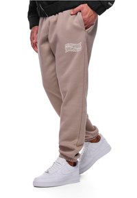 Beige bomulds-sweatpants med elastiske manchetter og sidelommer, med hvide grafikker på venstre ben og parret med hvide sneakers.