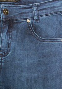 Jeans de mezclilla azul oscuro con una textura suave, diseño de cinco bolsillos, costuras visibles y acentos de metal en la cintura y los bolsillos.