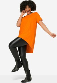 Kirkas oranssi ylisuuri t-paita lyhyillä hihoilla, yhdistettynä kiiltäviin mustiin leggings-housuihin ja niittikuvioisiin mustiin nilkkabootseihin. Silkkinen kangas.