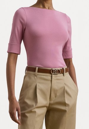 Femme portant un haut rose ajusté avec des manches au coude et un pantalon plissé beige, accessoirisée d'une ceinture marron avec une boucle dorée.