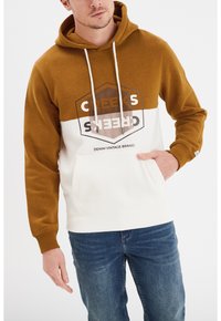Sudadera con capucha que presenta una sección superior marrón y una sección inferior blanca, con un bolsillo delantero y un diseño gráfico en negro. Se llevan jeans azules.