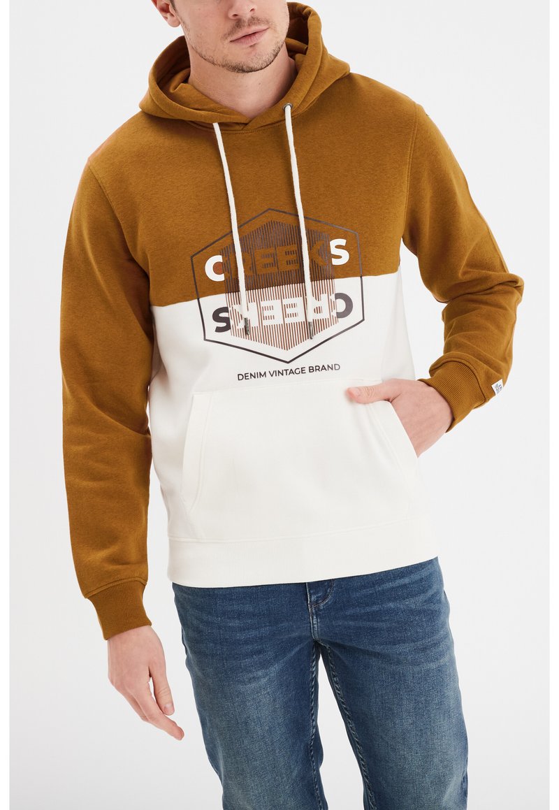 Sudadera con capucha que presenta una sección superior marrón y una sección inferior blanca, con un bolsillo delantero y un diseño gráfico en negro. Se llevan jeans azules.